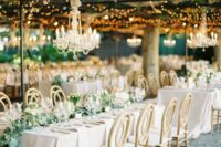 a neutral sophisticated wedding reception with neutral linens and chairs, with a greenery runner and a crystal chandelier over it