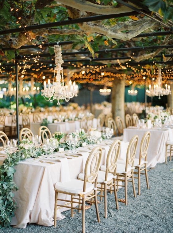 a neutral sophisticated wedding reception with neutral linens and chairs, with a greenery runner and a crystal chandelier over it
