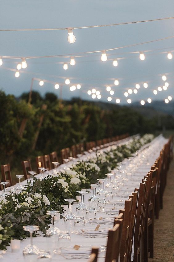 a neutral vineyard wedding reception with a greenery runner and white blooms, white linens plus string lights over the space