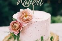 a pink buttercream speckle wedding cake with pink blooms, greenery and a gold calligraphy topper