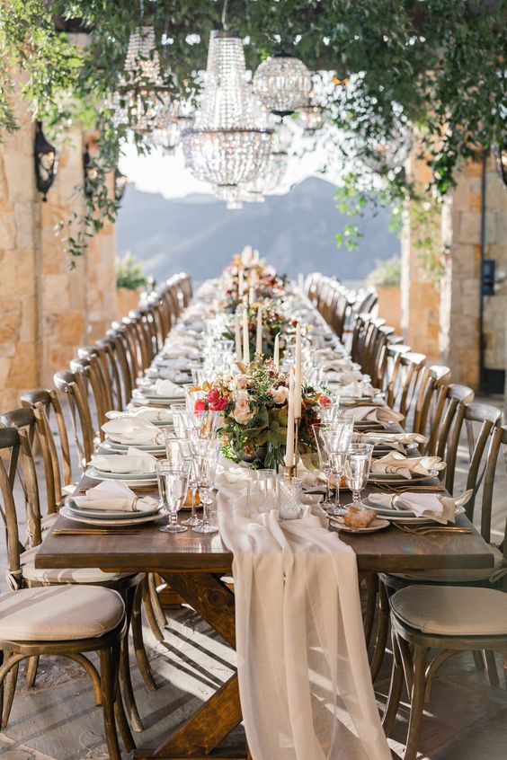 a refined vineyard wedding table setting with a neutral table runner, pink and coral blooms and greenery and crystal chandeliers over the space