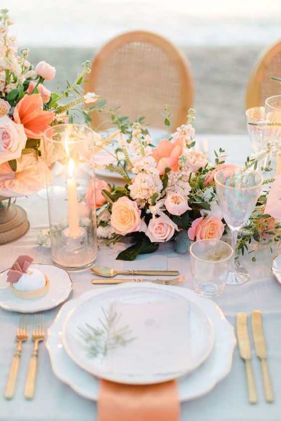 a romantic peach and blush wedding tablescape with blush and peachy blooms, orange napkins, gold cutlery and greenery plus tall candles