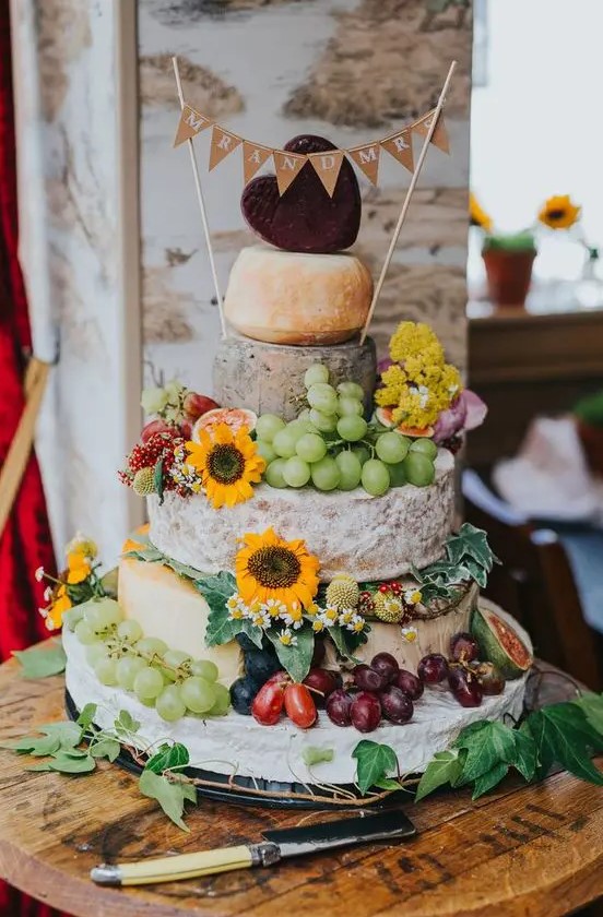a rustic cheese wheel wedding cake topped with super bold blooms, grapes, figs and tomatoes plus a burlap banner on top