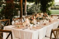a small and chic neutral vineyard wedding reception with neural blooms and greenery, pillar candles, neutral linens and chairs and a number of bulbs over the table