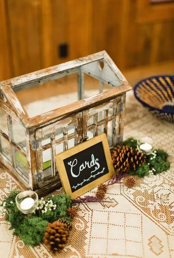 a small shabby chic glass house-style box for wedding cards decorated with a mini sign, pinecones, moss and candles