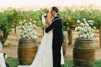 a vineyard wedding ceremony in the vines, with barrels showing off white florals and white petals on the ground