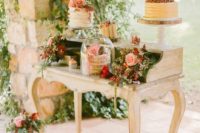 a vintage dessert table decorated with greenery, pink and red flowers and chic stands and a cloche