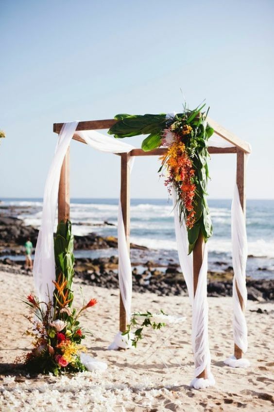a wedding arbor with white curtains, greenery, yellow and orange blooms with a sea view is a gorgeous idea for a beach wedding