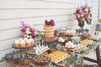 a wedding dessert table of a rustic weathered wooden table decorated with eucalyptus and bright flowers and delicious desserts