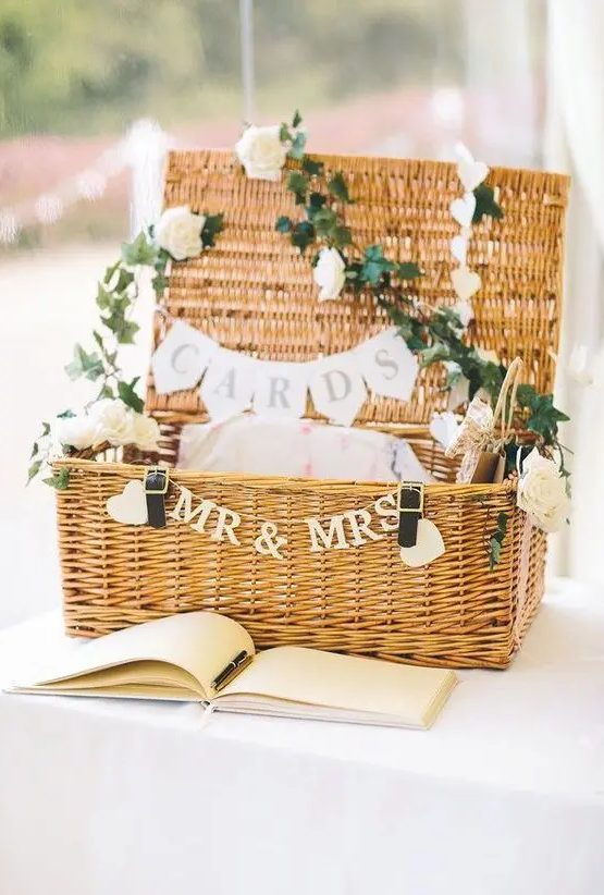 a woven suitcase decorated with greenery, white blooms and banners is a gorgeous idea for a rustic or just relaxed wedding