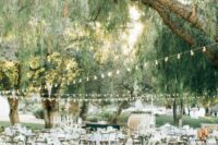 an elegant rustic wedding reception space with white tablescape and white blooms, some greenery and string lights over the space