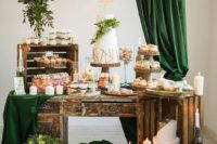 an elegant wedding dessert table of crates, lots of candles, a frame with greenery and dark green curtains and bulbs