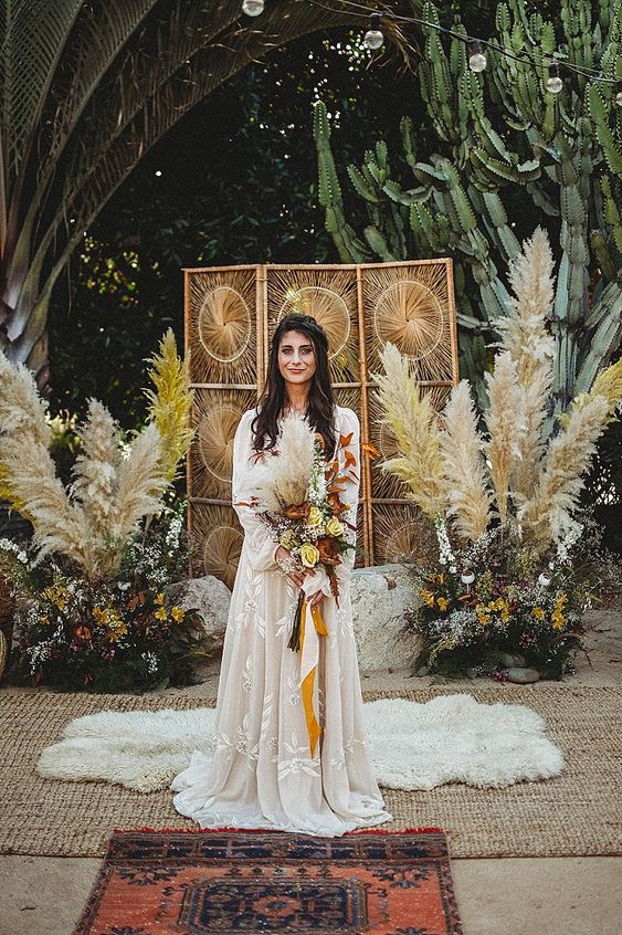 a 70s boho wedding backdrop with a woven screen, bright blooms, greenery, pampas grass and layered rugs