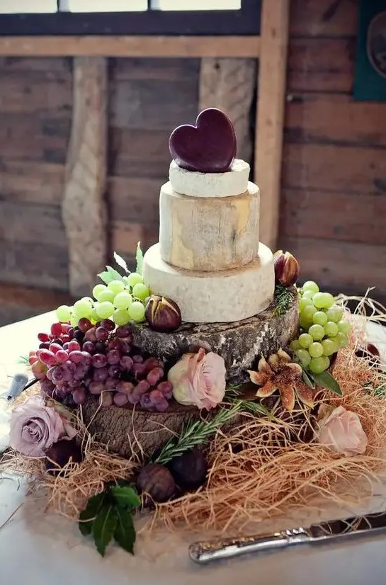 a beautiful cheese tower with grapes and figs, a cheddar heart on top displayed on wood stumps, with hay and roses for a rustic wedding