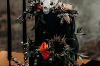 a black wedding cake with bold blooms, greenery and dried grasses, a skull on top is a bold and catchy idea for a Halloween wedding