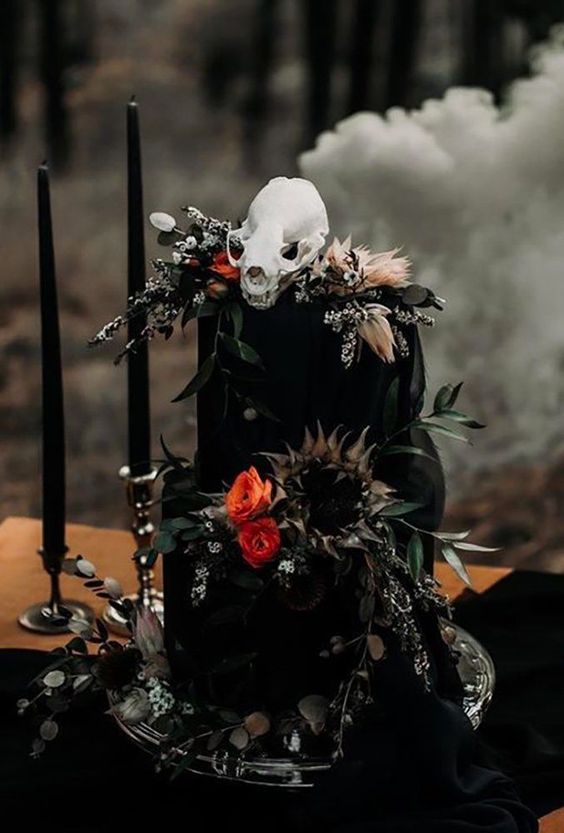 a black wedding cake with bold blooms, greenery and dried grasses, a skull on top is a bold and catchy idea for a Halloween wedding