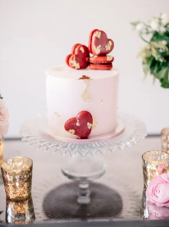 a blush wedding cake decorated with gold leaf and red heart shaped macarons is a cool idea for a Valentine wedding