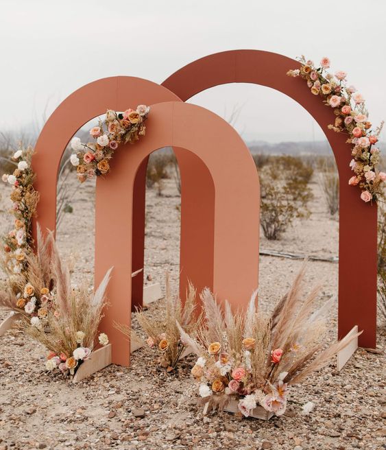 a bold fall wedding backdrop composed of three fall-colored arches, with neutral and pastel blooms and pampas grass for a boho wedding