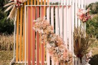 a bold fall wedding backdrop of mustard, pink and white planked pieces, pastel and neutral blooms, pampas grass and fronds is cool for a boho celebration