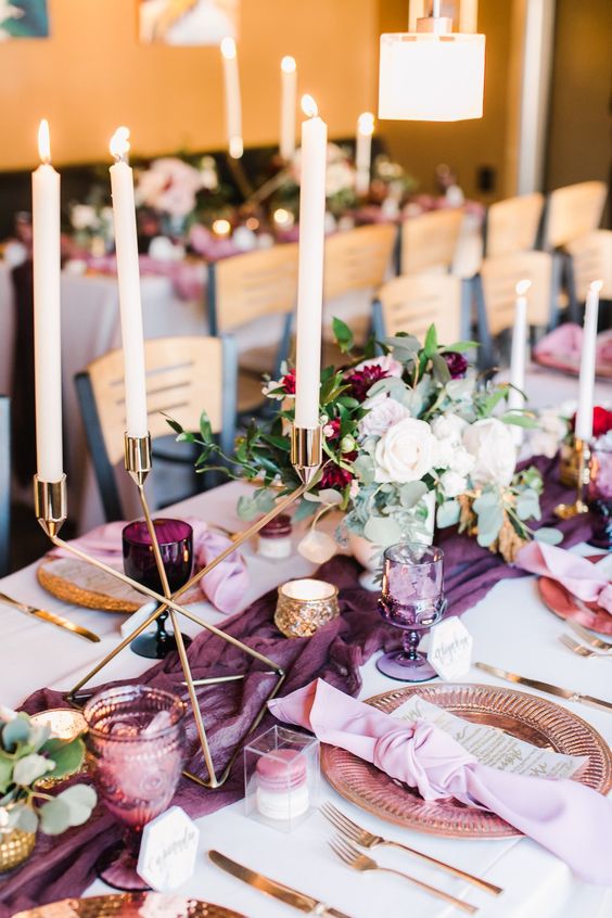 a catchy and lovely wedding table setting with a plum table runner and glasses, neutral blooms and greenery, copper chargers and candleholders