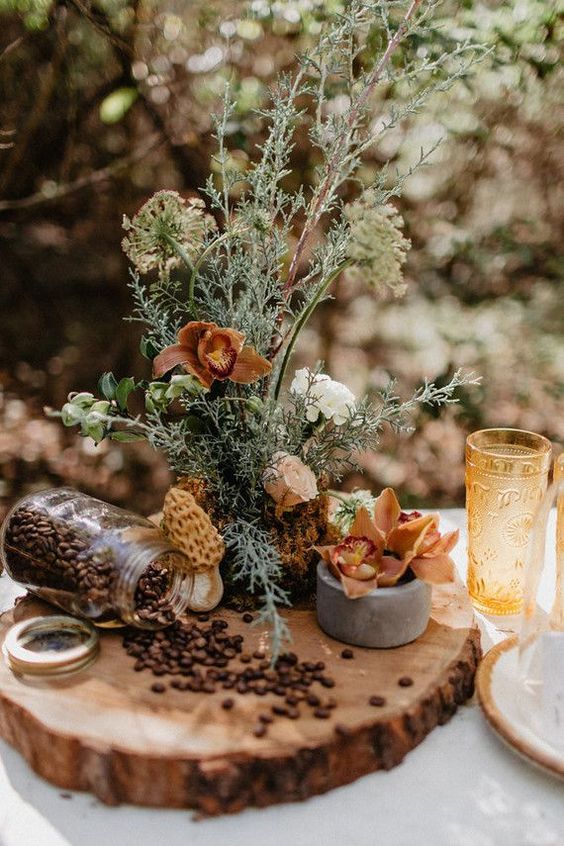 a catchy cluster fall wedding centerpiece of textural greenery, bold blooms, coffee beans on a wood slice and yellow glasses