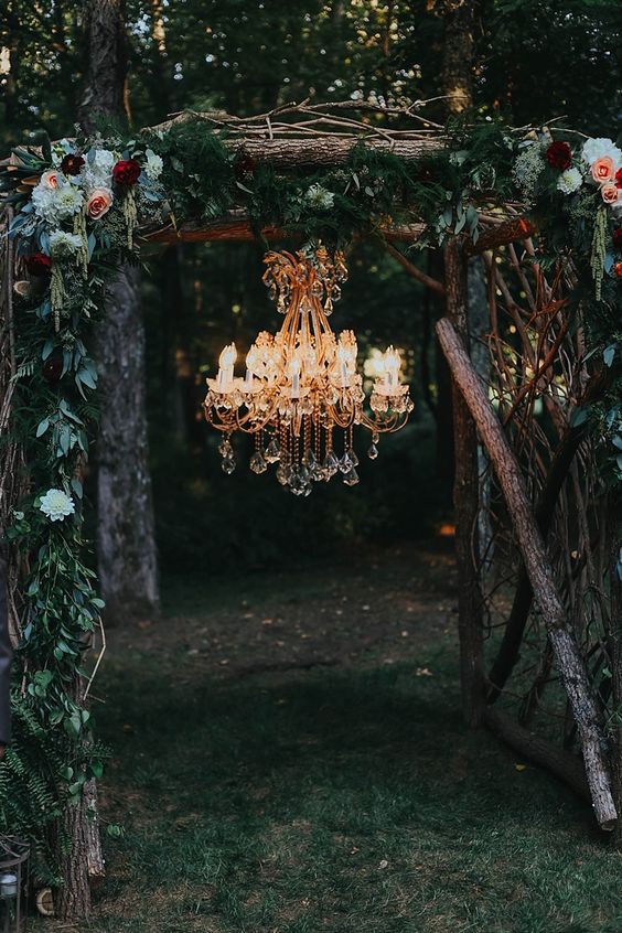 a chic fall woodland wedding arch of branches and logs, with plenty of greenery, neutral and burgundy blooms and a crystal chandelier