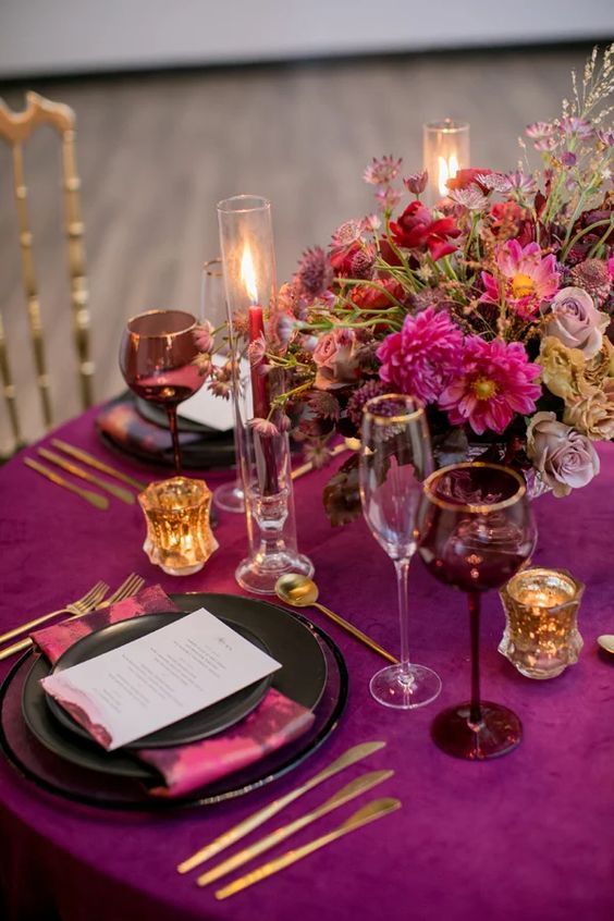 a chic plum-colored tablescape with pink blooms and napkins, gold cutlery and candleholders, tall and thin candles