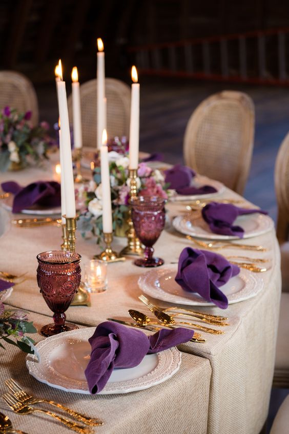 a chic wedding tablescape with neutral tablecloths, plum-colored napkins and glasses, tall candles and gold cutlery