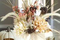 a fall wedding centerpiece of a white pumpkin, faux neutral blooms, dried grasses and branches and some pinecones and berries