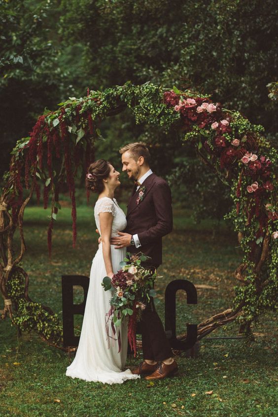 a fall woodland wedding arch of branches, with pink and burgundy blooms and lush greenery plus monograms is amazing