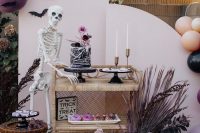 a gorgeous Halloween sweets table with a whimsy black cake with pink blooms and pink glazed donuts served by a skeleton