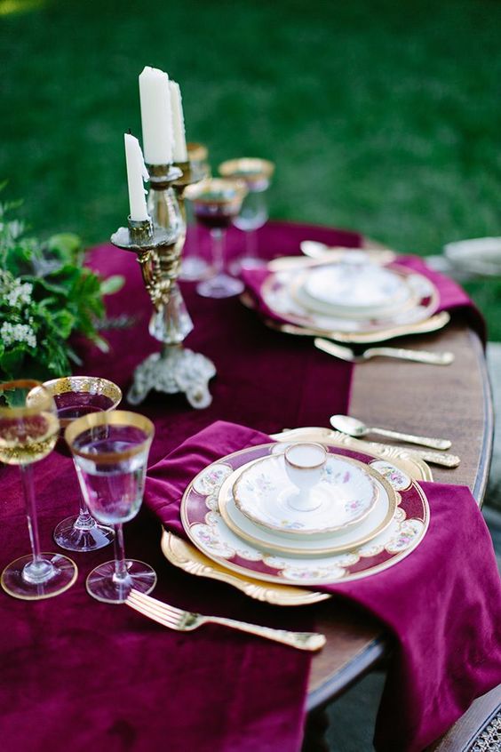 A gorgeous fall wedding tablescape with plum colored velvet textiles, greenery, tall and thin candles and gold rimmed glasses