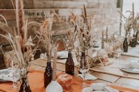 a lovely rustic Thanksgiving tablescape with orange runners, dark glass bottles with dried blooms and grasses and candles