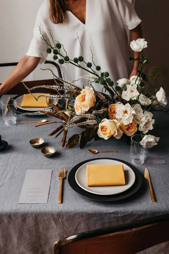 a refined modern Thanksgiving tablescape with a grey linen tablecloth, black and white plates, yellow napkins, a bold dimensional floral arrangement