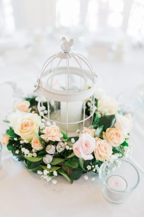 a refined wedding centerpiece with a white cage with a bird on top and candle inside, surrounded with greenery and pink and peachy blooms