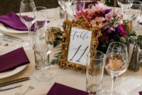 a refined wedding tablescape with a bold plum and pink centerpiece and plum-colored napkins, a table number in a frame is chic