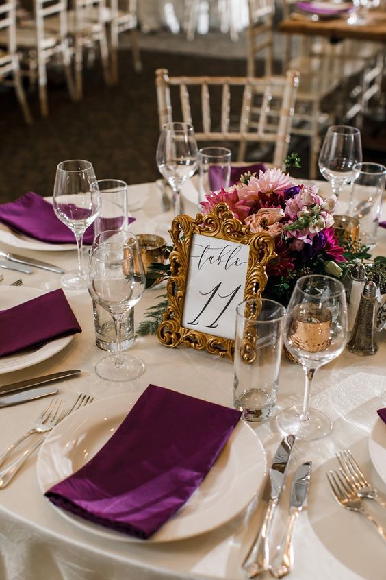 a refined wedding tablescape with a bold plum and pink centerpiece and plum-colored napkins, a table number in a frame is chic