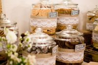 a rustic dessert table with large jars covered in burlap and lace, with neutral blooms and greenery and paper napkins