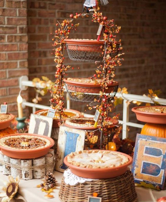 a rustic fall dessert table with a metal pie stand decorated with berries and leaves, wood and wicker stands, letters, pinecones and acorns