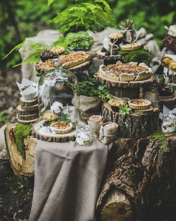 a rustic woodland dessert table with neutral linens, tree stumps and tree slices, greenery in pots and moss is adorable