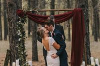 a simple and elegant fall woodland wedding arch with burgundy fabric, candles on tree stumps and greenery is amazing