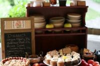a small and cute rustic dessert table with a wooden stand with plates, cookies and jars, pies and cupcakes on stands and a chalkboard