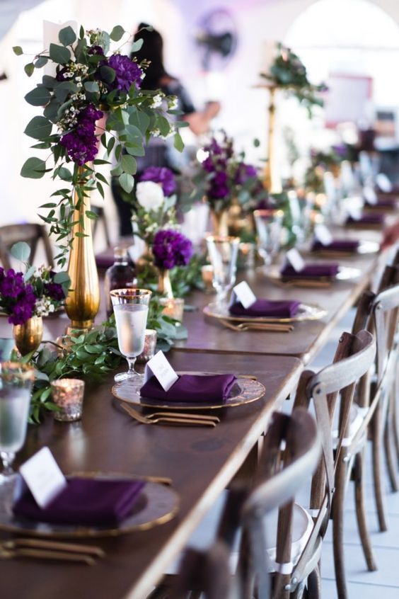 A sophisticated wedding tablescape with plum colored blooms and napkins, greenery and greenery runners, gold rimmed glasses