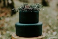 a stylish moody wedding cake with black and gold tiers, greenery on top and little white blooms on top is a lovely woodland wedding idea