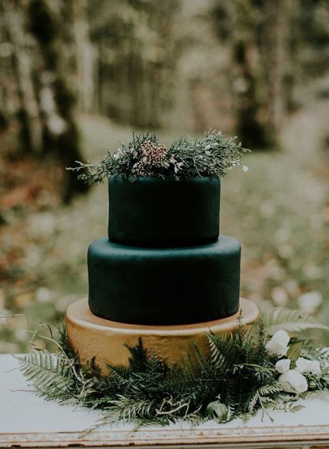 a stylish moody wedding cake with black and gold tiers, greenery on top and little white blooms on top is a lovely woodland wedding idea