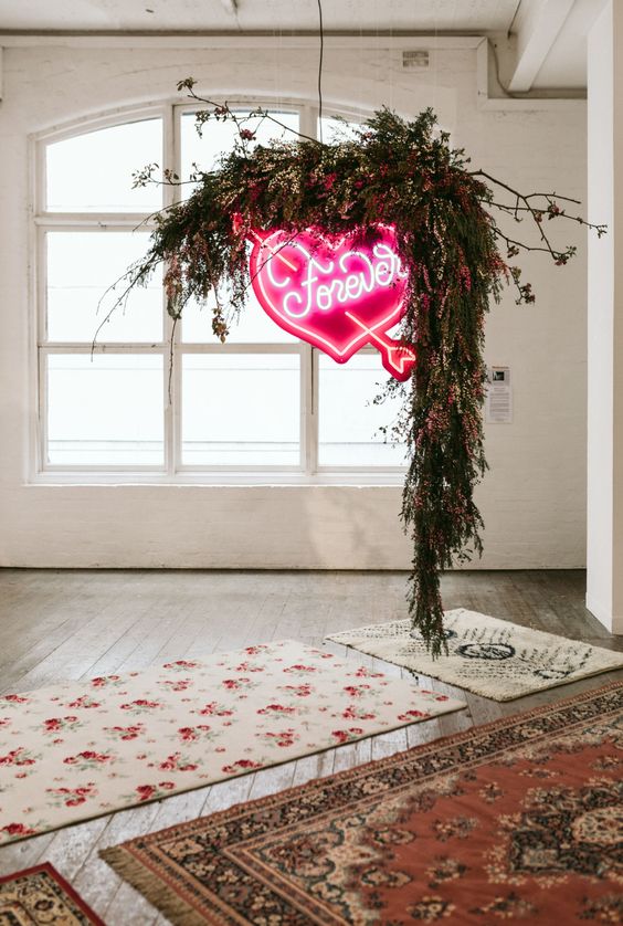 a wedding ceremony space with a bold rug, greenery and red blooms and a bold neon heart sign hanging is cool