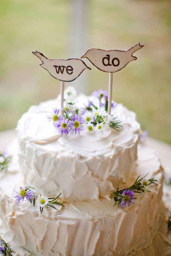 a white textural wedding cake decorated with purple and white blooms and topped with plywood bird-shaped cake toppers