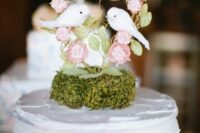 a white wedding cake with moss, a mini cake topper with a tree with two love birds is a cute idea for a wedding