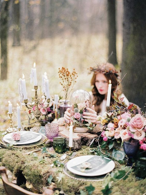 an echanted forest wedding reception table with a moss tablecloth and greenery, pink blooms and dried leaves, candles and colored glasses
