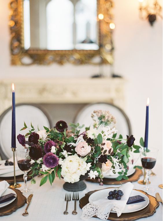 an elegant wedding tablescape with a white, blush and plum floral centerpiece, violet candles, gold chargers and blueberries